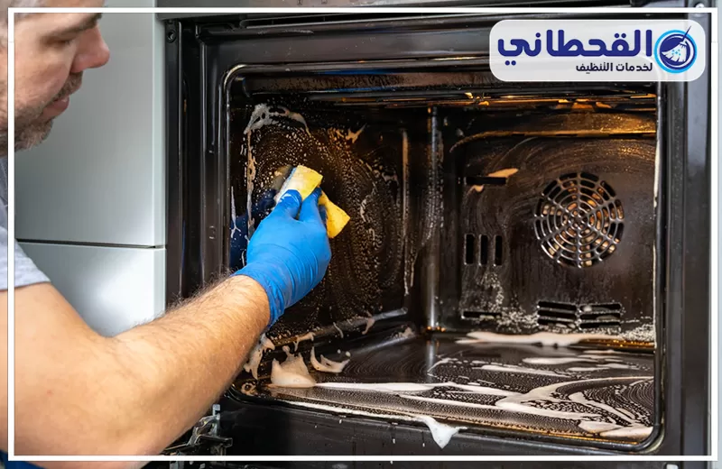 تنظيف أفران الغاز بسيهات 0548145142 شركة القحطاني يد ترتدي قفازًا أزرق تنظف داخل فرن كهربائي بفرشاة مع رغوة تنظيف كثيفة وإزالة دهون متراكمة على الجدران الداخلية مع ظهور شخص على الجانب أثناء التنظيف.