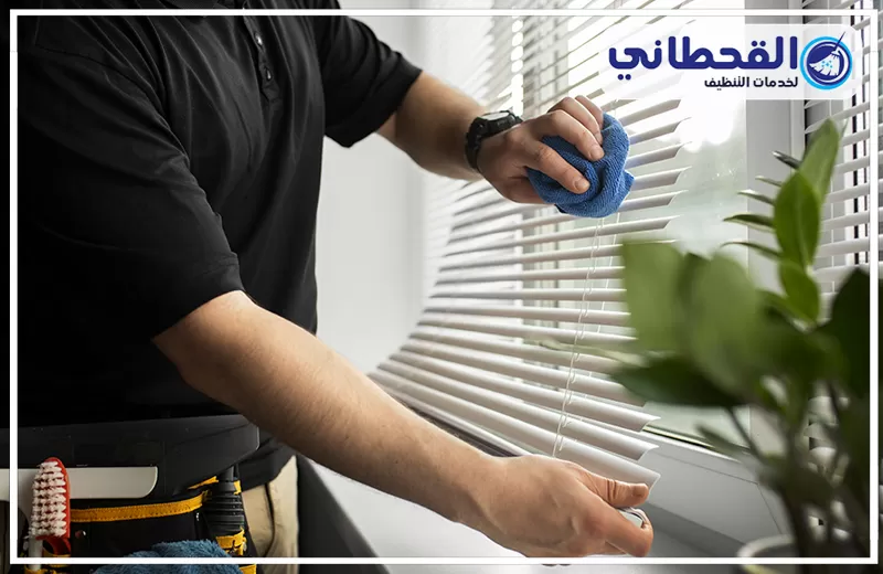 شركة تنظيف ستائر بعنك 0548145142 القحطاني عامل ينظف ستائر معدنية داخلية بقطعة قماش، مع ظهور شعار القحطاني لخدمات التنظيف.