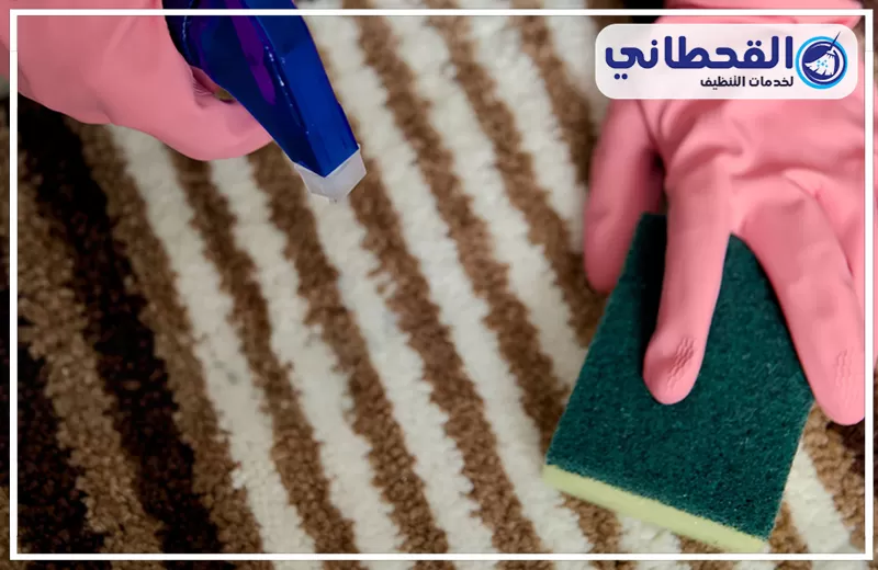 تنظيف السجاد قبل المناسبات والزيارات بصفوى 0548145142 شركة القحطاني. يدان ترتديان قفازات وردية تنظفان سجادة مخططة باللونين الأبيض والبني باستخدام بخاخ أزرق وإسفنجة خضراء، مع شعار القحطاني أعلى الصورة.
