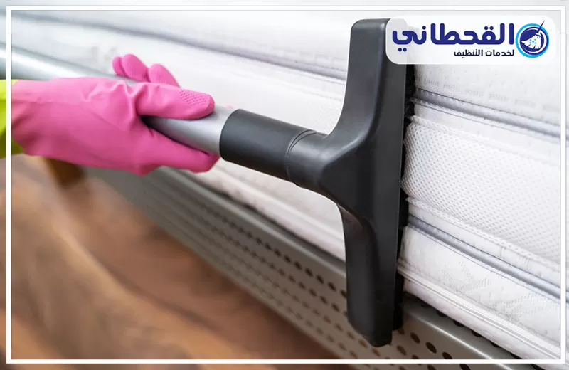 تنظيف فرشات السرير في رأس تنورة 0548145142 شركة القحطاني. يد بقفاز وردي تستخدم رأس مكنسة سوداء لتنظيف جانب مرتبة سرير بيضاء، مع ظهور قاعدة السرير الخشبية وشعار القحطاني أعلى الصورة.
