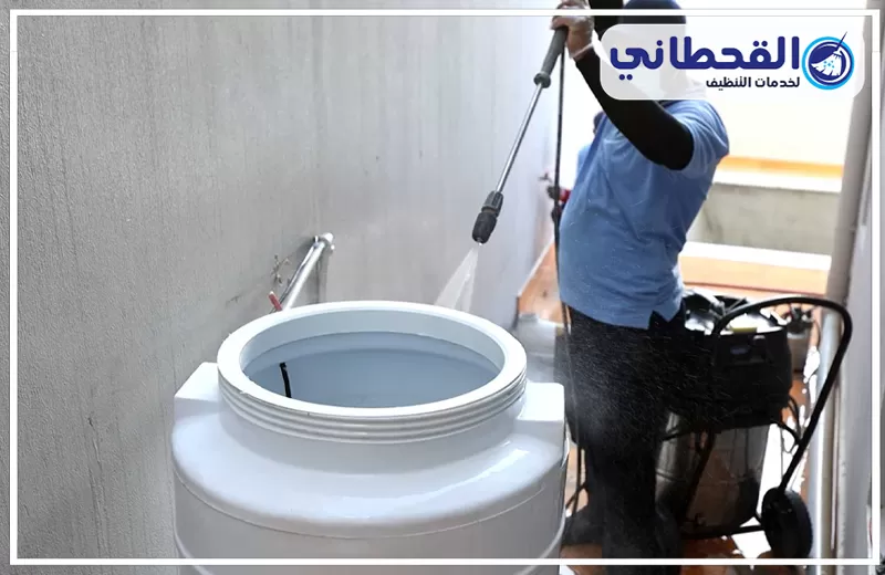 تنظيف خزانات العمائر برأس تنورة 0548145142 شركة القحطاني. فني يستخدم مضخة رش لتنظيف خزان مياه أبيض مفتوح، مع ظهور خرطوم ومعدة تنظيف بجواره وشعار القحطاني أعلى الصورة.