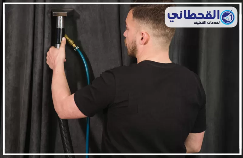 تنظيف ستائر مجالس وغرف نوم برأس تنورة 0548145142 شركة القحطاني. عامل ينظف ستائر سوداء بجهاز شفط وخرطوم أزرق مع شعار القحطاني أعلى الصورة
