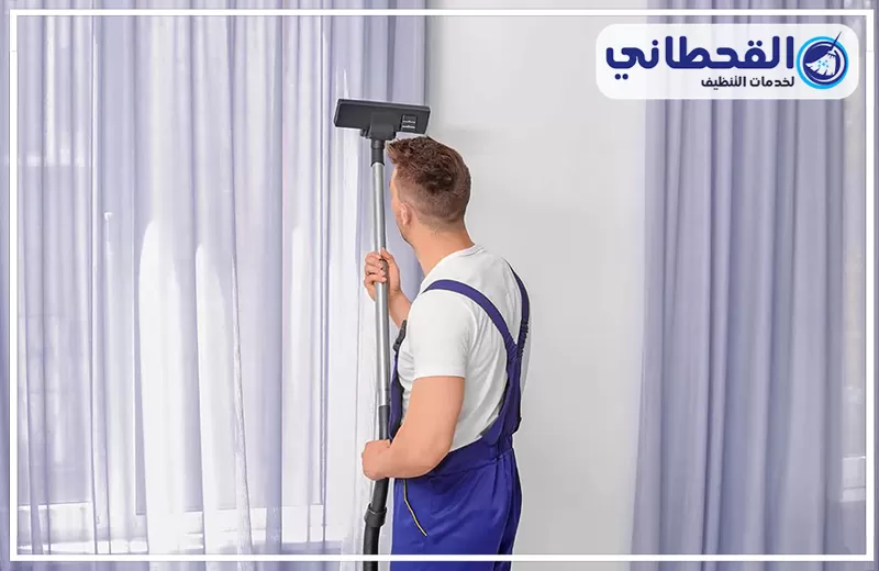تنظيف ستائر رول وبلاك أوت بصفوى 0548145142 شركة القحطاني. فني بزي أزرق يستخدم مكنسة طويلة لتنظيف ستائر رمادية معلقة أمام نافذة، مع ظهور الجدار الأبيض وشعار القحطاني أعلى الصورة.