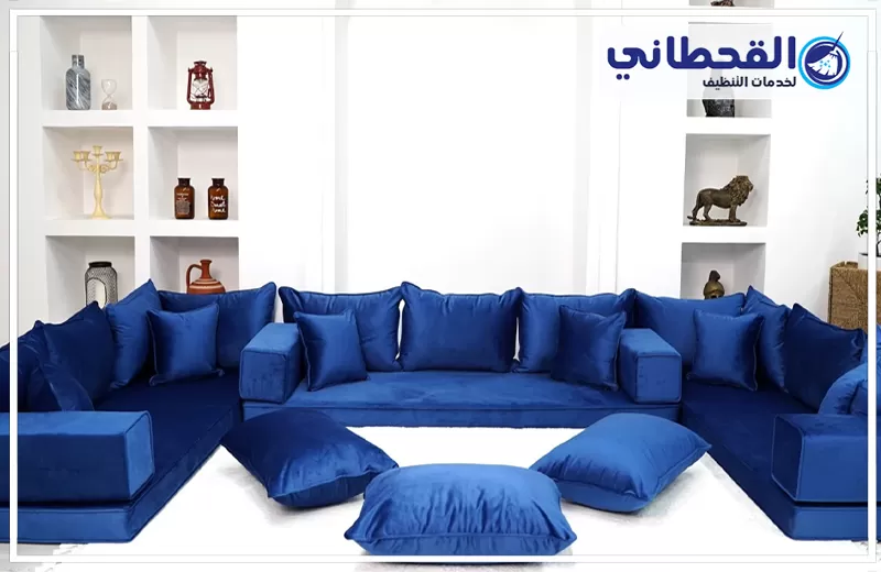 تنظيف مجالس بعد المناسبات بعنك 0548145142 شركة القحطاني. مجلس كنب أزرق كبير على شكل حرف U داخل غرفة بيضاء، يحتوي على مساند ظهر ووسائد زرقاء متعددة ووسائد أرضية أمامية، مع رفوف ديكور جانبية وشعار القحطاني أعلى الصورة.