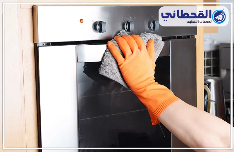 تنظيف افران آمن على الطعام بعنك 0548145142 شركة القحطاني يد ترتدي قفازًا برتقاليًا تمسح واجهة فرن استيل بقطعة قماش، مع إبراز تنظيف الأفران وتلميع السطح الخارجي.