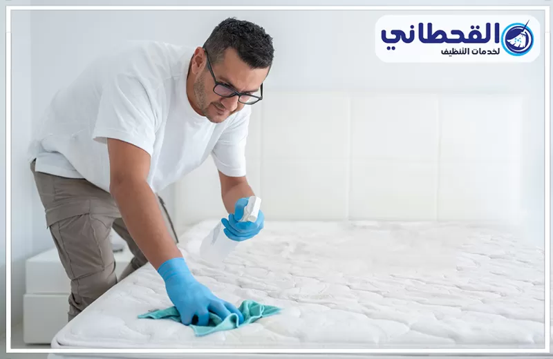 اسعار شركة تنظيف مراتب السرير بالخبر 0548145142 شركة القحطاني عامل تنظيف يرتدي قفازات ويستخدم بخاخًا وقطعة قماش لتنظيف مرتبة سرير، مع شعار القحطاني لخدمات التنظيف.