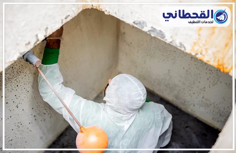تنظيف خزانات الفيبر جلاس برأس تنورة 0548145142 شركة القحطاني. فني ببدلة حماية بيضاء داخل خزان مياه خرساني، يمسك أداة تنظيف، مع ظهور بقع على الجدران وفتحة الخزان من الأعلى وشعار القحطاني.