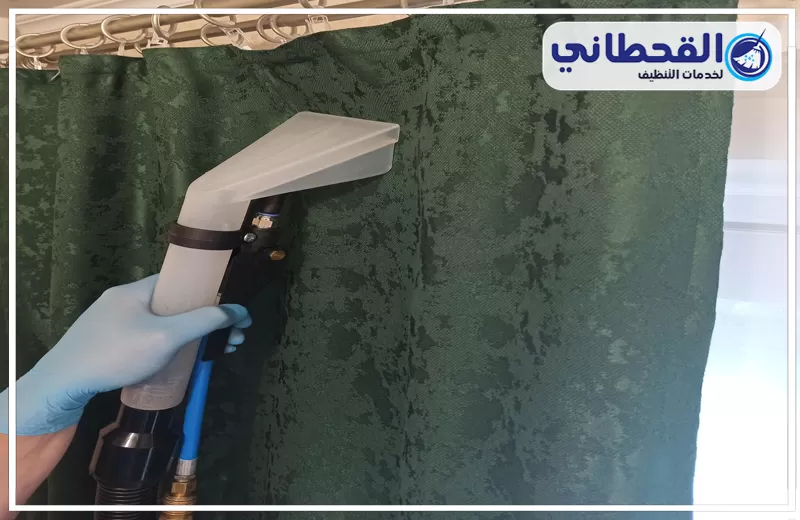 إزالة بقع الستائر بصفوى 0548145142 شركة القحطاني. يد بقفاز أزرق تستخدم جهاز تنظيف بالبخار على ستارة خضراء معلقة بحلقات معدنية، مع ظهور آثار بلل وشعار القحطاني أعلى الصورة.