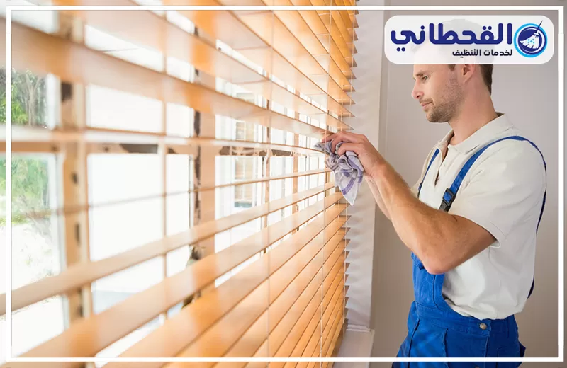 غسيل ستائر في مكانها بصفوى 0548145142 شركة القحطاني. فني يرتدي زيًا أزرق ينظف ستائر خشبية معلقة بجوار النافذة بقطعة قماش، مع ظهور شرائح الستارة وشعار القحطاني أعلى الصورة.