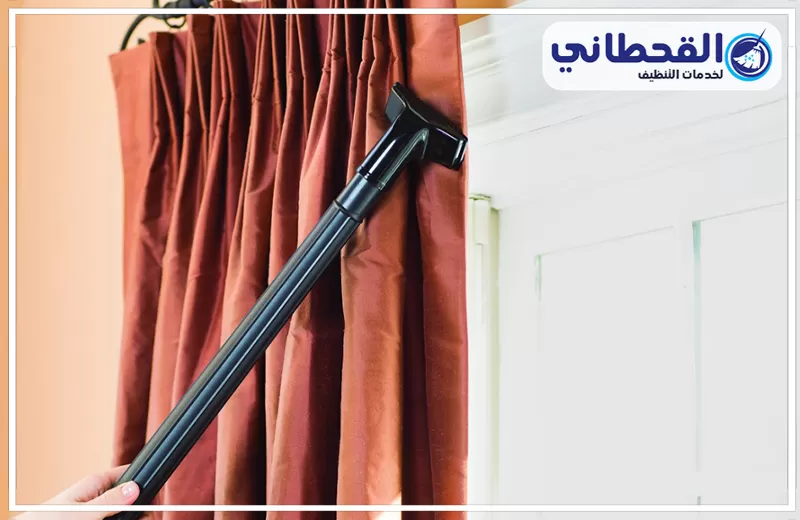 تنظيف ستائر بالبخار بصفوى 0548145142 شركة القحطاني. يد تستخدم رأس مكنسة سوداء لتنظيف ستارة بنية معلقة بجوار باب أبيض، مع ظهور طيات الستارة وشعار القحطاني أعلى الصورة.