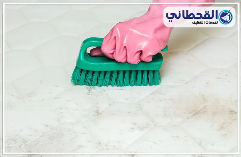 تعقيم مراتب السرير في رأس تنورة 0548145142 شركة القحطاني. يد ترتدي قفازًا ورديًا تنظف مرتبة سرير بيضاء بفرشاة خضراء، مع ظهور بقع خفيفة على المرتبة وشعار القحطاني أعلى الصورة.