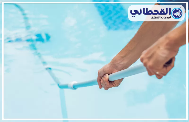 تنظيف وتعقيم مسابح بالظهران 0548145142 شركة القحطاني. يد تمسك عصا تنظيف داخل مسبح بمياه زرقاء فاتحة، مع ظهور أداة تنظيف أسفل الماء وتموجات خفيفة على السطح.