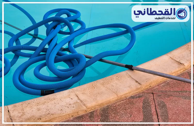 تنظيف فلتر المسبح بالظهران 0548145142 شركة القحطاني. خرطوم تنظيف أزرق ملتف داخل مسبح بمياه صافية، مع عصا تنظيف معدنية ممتدة على حافة المسبح.
