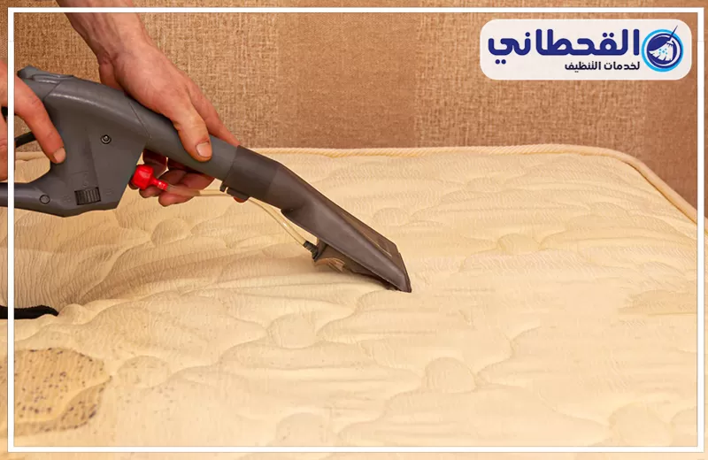غسل مفارش السرير في المنزل برأس التنورة 0548145142 شركة القحطاني. يد تمسك جهاز تنظيف رمادي فوق مرتبة سرير بيج مبطنة، مع ظهور قماش بني بالخلفية وشعار القحطاني أعلى الصورة.