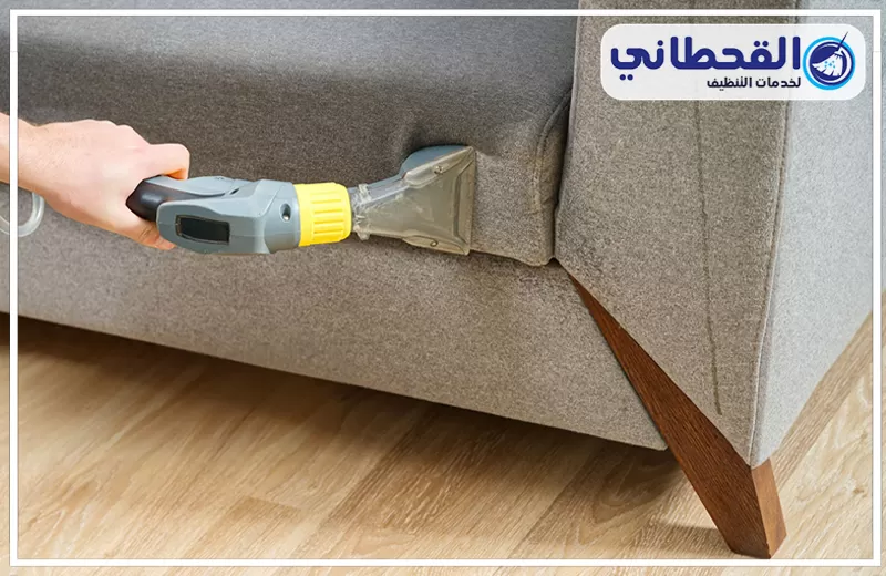 تنظيف كنب قماش براس تنورة 0548145142 شركة القحطاني. يد تستخدم جهاز شفط مخصص لتنظيف الكنب داخل شقوق كنبة قماش رمادي مع توجيه الأداة بين الوسائد وحافة الذراع فوق أرضية خشبية.