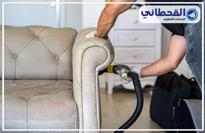 تنظيف كنب جلد في راس تنورة 0548145142 شركة القحطاني. يد عامل تنظيف يرتدي قفازات بيضاء يستخدم خرطوم جهاز شفط لتنظيف ذراع كنبة قماش بيج داخل غرفة.