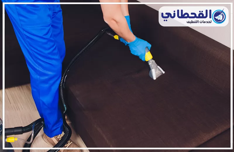 تنظيف كنب بالبخار براس تنورة 0548145142 شركة القحطاني. عامل يرتدي قفازًا أزرق وبنطال عمل أزرق يستخدم فوهة جهاز شفط لتنظيف سطح كنبة قماش بني مع وجود خرطوم الجهاز على الأرضية الخشبية.